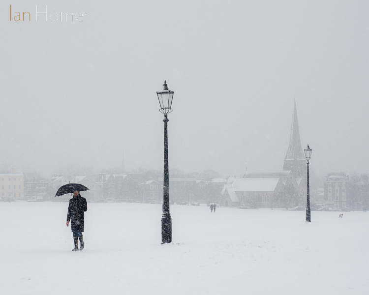 Blackheath Common now white with heavy snow falling. A man strolls holding an umbrella to shield himself from the flakes.