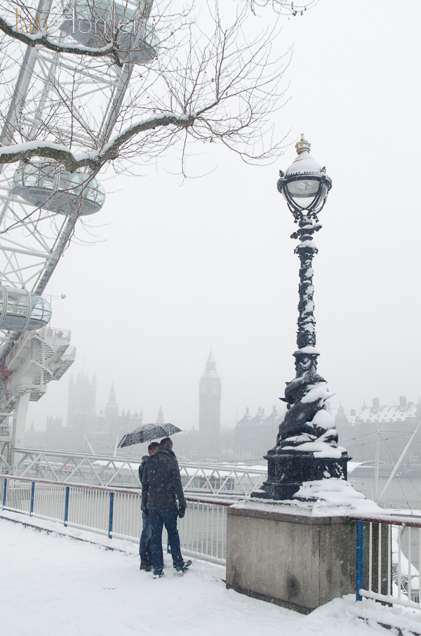 South Bank; Eye and Palace of Westminster under snow, two figures talk, sheltered by their umbrella.