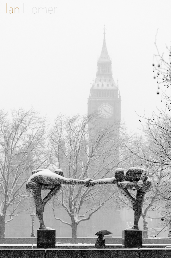 Division Bell: The sporting sculpture by Rick Kirby tensely looms over an umbrella carrying walker's head with the clock-tower housing Big Ben almost seemingly looking on.